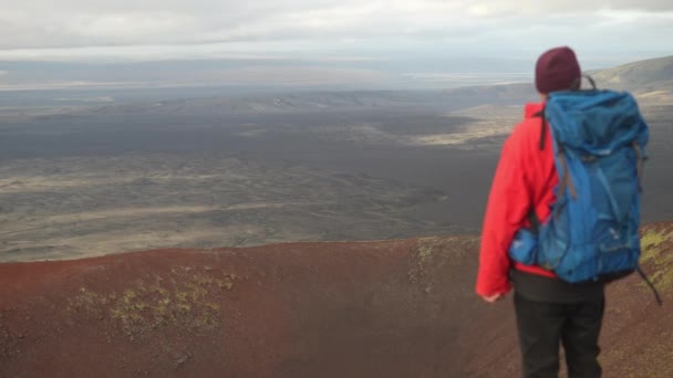 バックビューショットは アイスランドのラウダスカルクレーターに立っているジャケットとバックパックを着た男をキャプチャし カメラは赤い火山の土にズームします — ストック動画