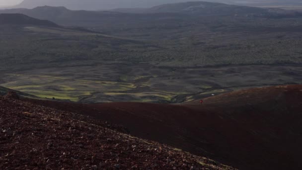 ラウダスカルクレーターの赤と砂の地形を暗い火山の風景の背景にハイキングする男の広いショット — ストック動画
