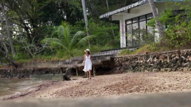 Beyaz elbiseli bir bayanın gündüz vakti Tayland 'da Ko Chang' de terk edilmiş bir evin yanında huzur içinde yürüdüğü geniş bir fotoğraf.