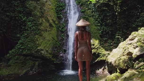 後ろから見たビキニとわら帽子の女性は 彼女の周囲を確認するために回る前に滝を賞賛 — ストック動画