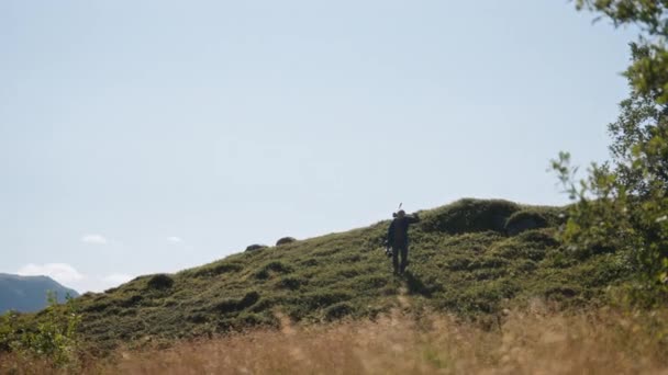 映画製作者は緑色の草を横切って歩き 青空の背景にカメラと三脚を運びます — ストック動画