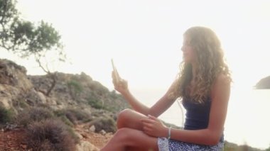 Mavi bluzlu bir kız, parlak güneşin altında doğada akıllı bir telefonla selfie çekiyor.