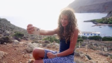Mavi bluzlu ve etekli bir kız, güzel dağ manzaralı fotoğraflar çekiyor ve arka planda deniz, doğanın güzelliğiyle selfieleri birleştiriyor.