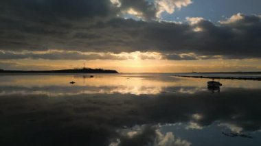 Alçaktan uçan bir dron sakin bir sahil boyunca süzülür, arka planda manzaralı günbatımını ve bulutlu gökyüzünü yakalar.