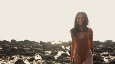 Kahverengi tek parça mayo giymiş bir kadın, gün batımında engebeli, kayalık sahilde yürürken saf neşe yaşar.