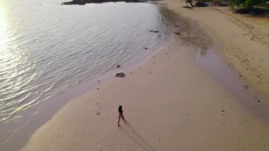 Büyüleyici bir hava perspektifinden bir kadın gün batımında Ko Mak Sahili 'nin el değmemiş kıyılarında yürüyor.