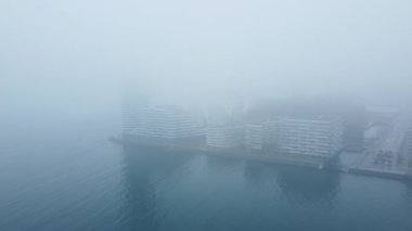 İHA tarafından çekilen hava görüntüsü, binaları ve gökdelenleri olan yoğun sisli bir limana yavaş yavaş yaklaştığını gösteriyor.