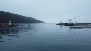 Alçaktan uçan bir dron, deniz kıyısındaki bir limanın üzerinde süzülür, sisli bir siluete karşı kurulmuş küçük teknelerin üzerinden geçer.
