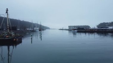 Alçaktan uçan bir dron suyun üzerinde süzülür, teknelerin üzerinden geçer ve sisli ufuk çizgisinin arka planına karşı yakınlardaki liman binalarını ortaya çıkarır.