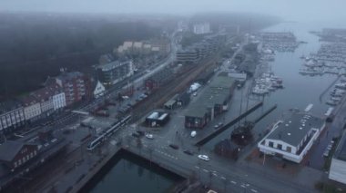 Bir İHA sisli bir şehir manzarası yakalar, limana demirlemiş tren, araba ve botları gösterir.