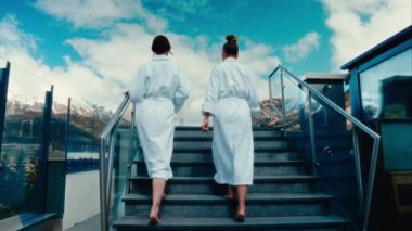 Beyaz mayo giymiş iki kadının, güzel bir dağ manzarasının zemininde havuza doğru yürürken çekilmiş yavaş çekimde çekilmiş bir dikiz görüntüsü.