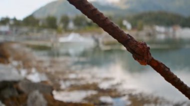 Ön planda paslı metal çubuğu olan, dağlık bir limana yanaşmış bulanık bir tekne görüntüsü.