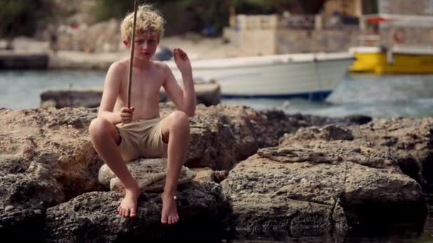 Ein Junge Sitzt Auf Einem Felsen Und Wirft Mit Einer — Stockvideo