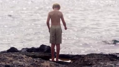 Arka perspektiften büyüleyici görüntüler, kayalık bir kıyı şeridinde balık tutan genç bir çocuğu gösteriyor.