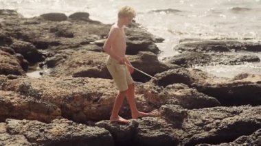 Genç bir çocuk elinde bir olta tutarak kayalık bir sahilin engebeli arazisinde dikkatle geziniyor.