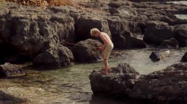 Kayalık bir sahilde bir ağaç dalından yapılmış geçici bir çubukla ustalıkla balık tutarken genç bir çocuğun becerisini gösteren büyüleyici görüntüler.