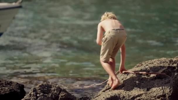 Boy Navigates Cautiously Rocky Shore Holding Fishing Stick Blurry Boat — Stock Video
