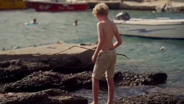 Joven Navegando Cautelosamente Por Costa Rocosa Con Telón Fondo Barcos — Vídeo de stock