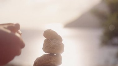 Ayrıntılı bir perspektif, huzurlu bir günbatımında sakin deniz manzarasına karşı kaya kuleleri inşa eden elleri gösteriyor.