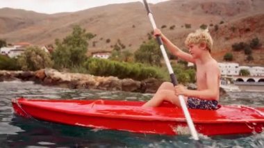 Genç bir çocuk Girit 'in manzaralı sularında parlak kırmızı bir kanoyla manevra yapıyor. Akdeniz manzaralarının hayranlık uyandıran güzellikleriyle çevrili.