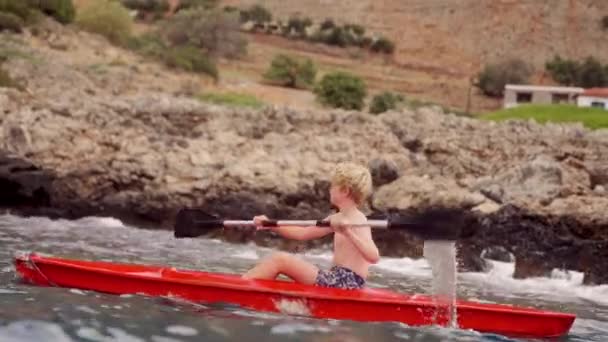 一个小男孩驾着一艘生机勃勃的红色皮划艇沿着克里特岛岩石海岸航行 — 图库视频影像