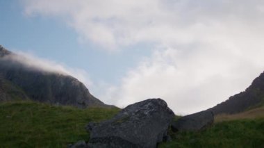 Sağdan sola doğru panoramik bir kamera engebeli dağ zirvelerinde sürüklenen bulutları gösterir.