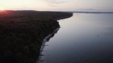 İnsansız hava aracı, gün batımında küçük bir tekne ile süslenmiş sakin sularla buluştuğu sahil ormanının üzerinde zarifçe süzülür.