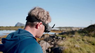 Açık mavi bir gökyüzü, dağ ve dingin bir göl üzerinde, mavi ceketli ve FPV gözlüklü bir film yapımcısı, yerden bir dronu yönetir.