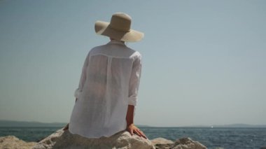Beyaz plaj gömleği ve hasır şapkalı bir kadının kayalık bir kıyıda otururken berrak mavi gökyüzünün altındaki sakin denizi seyrederken bir arka plan görüntüsü.