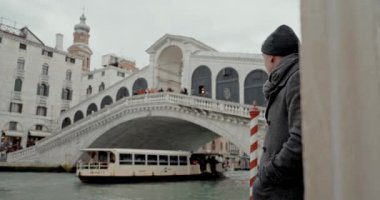 Siyah ceketli şık bir adam ikonik Rialto Köprüsü 'nün yanında duruyor ve altından zarif bir şekilde süzülüyor.
