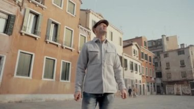 Gri bir şapka ve açık renk bir ceket giyen bir adam Venedik sokaklarında kendisini çevreleyen tarihi binalara hayranlıkla bakıyor.