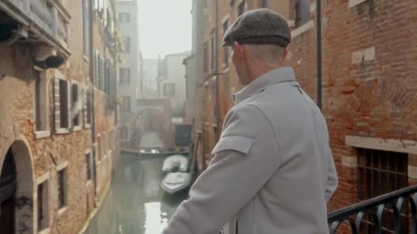 ヴェネツィアの歴史的建造物と運河を鑑賞しながら橋の上に立っている男の後ろからの眺め — ストック動画