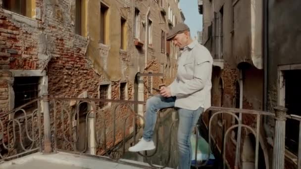 威尼斯运河附近一座小桥的金属栏杆上坐着一个戴着夹克和帽子的男人 他在光天化日之下拿着老式建筑的背景用手机发短信 — 图库视频影像