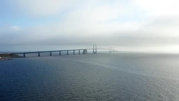 ドローンは穏やかな海の上の右側に優雅に光り輝き 遠くにある象徴的なグレートベルト橋の絶え間ない美しさを捉えています — ストック動画