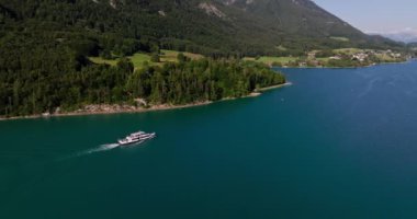 Avusturya gölünde seyreden bir teknenin insansız hava aracı görüntüsü. Etrafı yemyeşil dağların nefes kesici manzarasıyla çevrili.