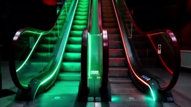 Yeşil, kırmızı ve sarı ışıklarla aydınlatılan bir çift yürüyen merdiven, renkli ışıkları yansıtan metalik basamaklar ve aydınlatma efektini artıran cam kenarlar.