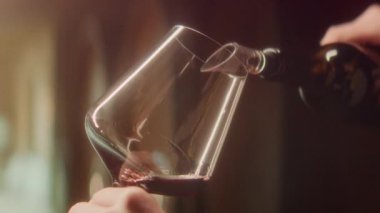 Kırmızı şarap şişesinden şarap bardağına dökülüyor. Arka planda sıcak tonlarda bulanıklık var.