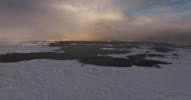 Bir dron kış manzarası üzerinde uçuyor, zemini karla kaplı, ortasında kısmen donmuş koyu renkli bir su kütlesi ve arka planda yoğun bulutların güzel bir görüntüsü var.