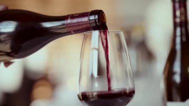 Siyah bir şişeden temiz bir şarap bardağına kırmızı şarap döken yakın plan bir el resmi, kapalı bir arka fona kurulmuş.