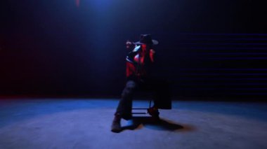 Bir el kamerası bir içeri bir dışarı zumluyor, siyah şapkalı bir kadını mavi ve kırmızı bir odada dans ederken yakalıyor.