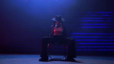 Bir el kamerası görüntüsü, siyah şapkalı yetenekli bir kadını parlak mavi ve kırmızı lazer ışıklarıyla loş bir odada sandalye dansı yaparken yakalıyor.