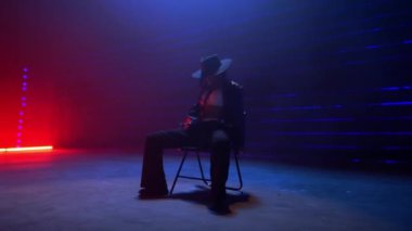 Siyah şapkalı yetenekli bir kadın tarafından çekilen büyüleyici bir sandalye dans gösterisi sol ve sağ zarifçe kıvrılarak loş bir odada canlı kırmızı ve mavi lazer ışıklarıyla çekiliyor.