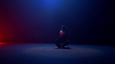 Tam statik bir çekim büyüleyici siyah bir elbise ve şapka giymiş bir kadını parlak kırmızı ve mavi lazer ışıklarıyla loş ışıklı bir odada büyüleyici bir sandalye dansı yaparken yakalar.