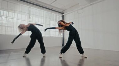 Sabit bir perspektiften çekilen, parlak siyah kıyafetler giymiş iki kadın büyüleyici bir koreografi sergiliyor.