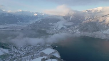 Bir dron evlerin üzerinde uçuyor ve yüzeyinde ince bir sis tabakası bulunan büyük bir göl yakınındaki karla kaplı yapılar, görkemli dağları ve mavi gökyüzünü görüyor.