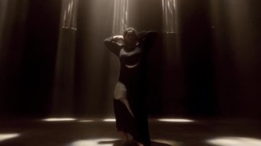 Stüdyo ışıklarının sıcacık parıltısıyla aydınlatılmış bir kadının zarif dansını gösteren kavisli bir el kamerası.