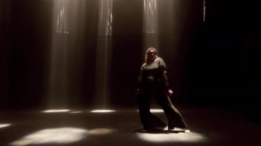 Sağa doğru hareket eden bir izleme çekimi bir kadının zarif hareketlerini gösteriyor. Stüdyo ışıklarının narin parıltısında.