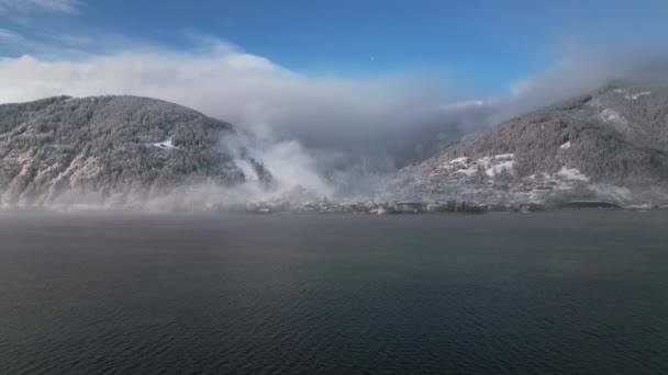 広大で穏やかな湖の景色は 雪に覆われた山のベースに小さな村を部分的に覆い 青い空の下に厚い霧で毛布に覆われたピークを間違えています ストック動画