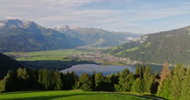 Bir hava manzarası, sakin bir gölün güzelliğini yakalar, çevresindeki manzarayı yansıtır, yemyeşil ormanların arasına yerleşir ve berrak bir gökyüzünün altında yükselen dağlar.