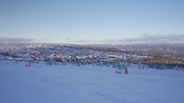 Bir kış manzarası, karla kaplı ağaçların arasında yuva yapmış küçük bir kasabayı barındırır. Ön planda el değmemiş geniş bir kar tabakası hakim olurken, arka planda dönen dağlar yer alıyor.